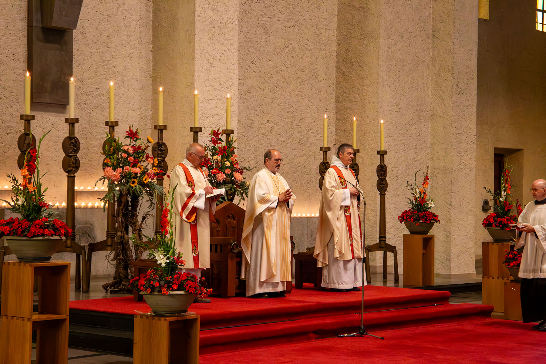 Drei Priester im weißen Gewandt umrahmt von Kerzen und Blumen