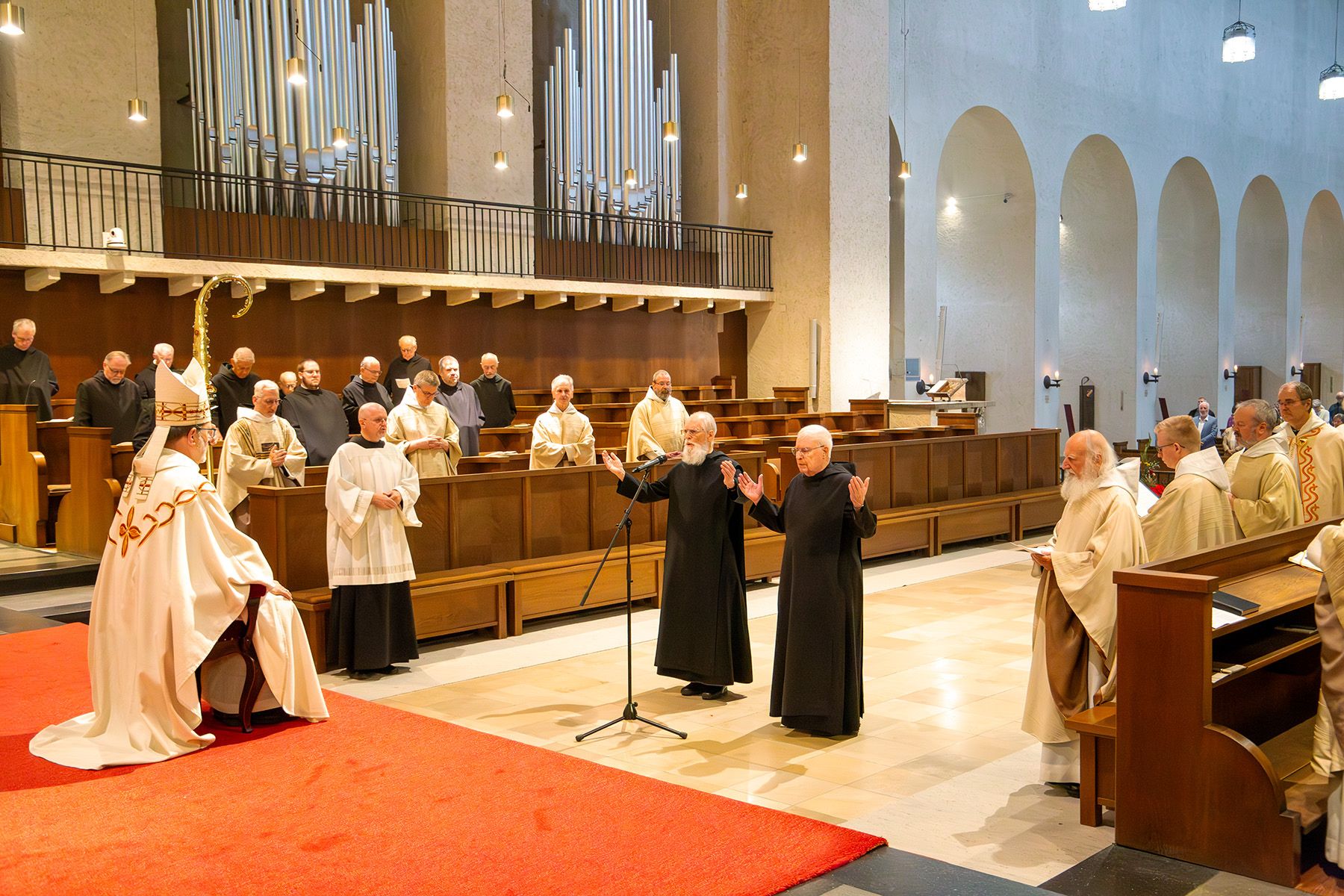 Zwei Mönche stehen mit erhobenen Händen in der Kirche vor einem Mann in weißem Gewandt und mit Abthut